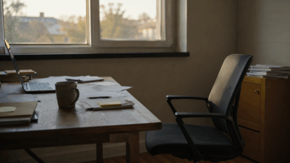 Ein leerer Bürostuhl steht zurückgeschoben an einem Schreibtisch mit Laptop, Papieren und Kaffeetasse. Das ruhige Bild im warmen Nachmittagslicht symbolisiert Überforderung, Burnout, innere Erschöpfung und den Wunsch nach Entlastung durch Hypnose gegen Stress.